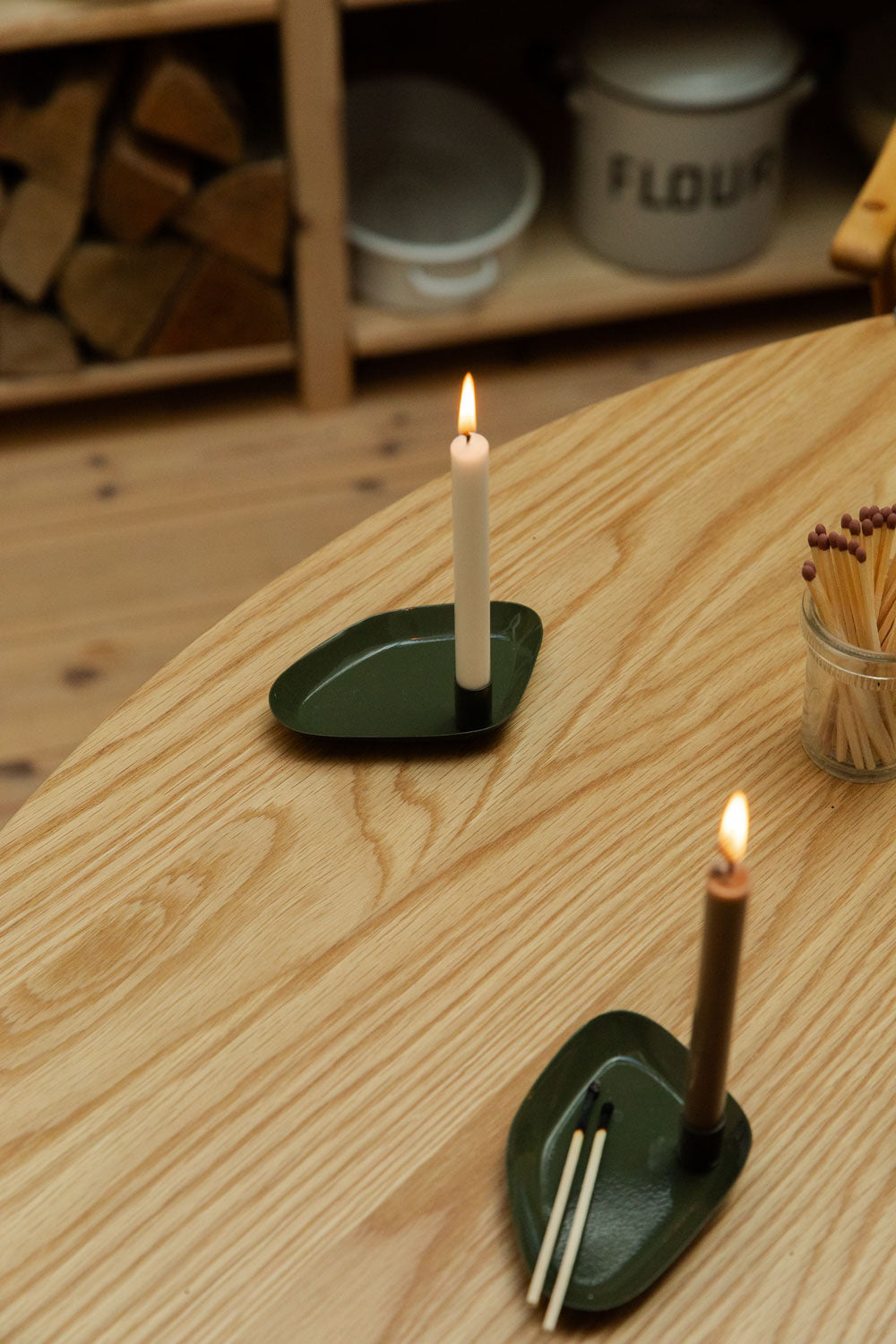 Two candle holders with lit candles on a wooden surface, with a shelf in the background.
