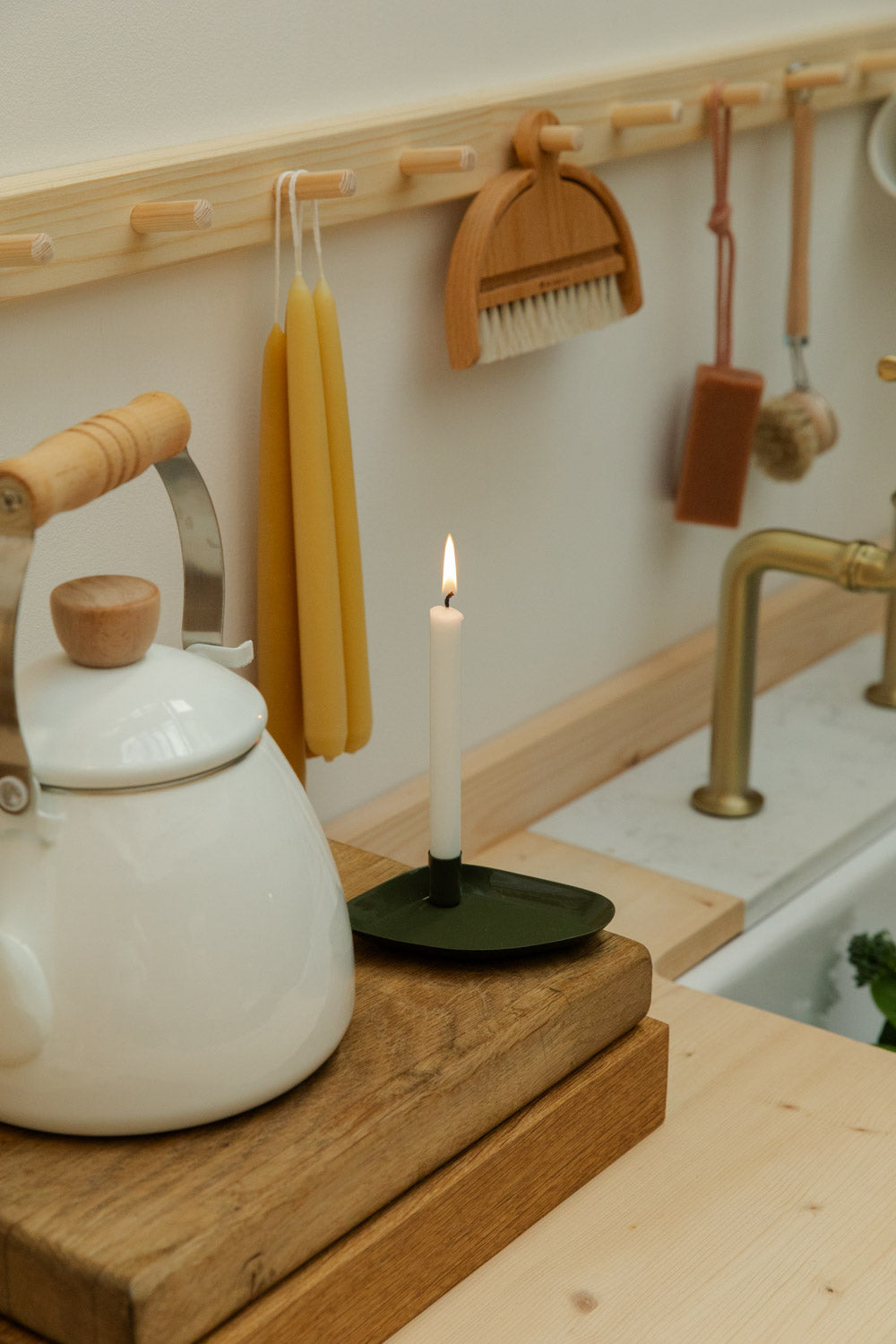 White teapot on a wooden cutting board with a lit candle, kitchen utensils in the background.
