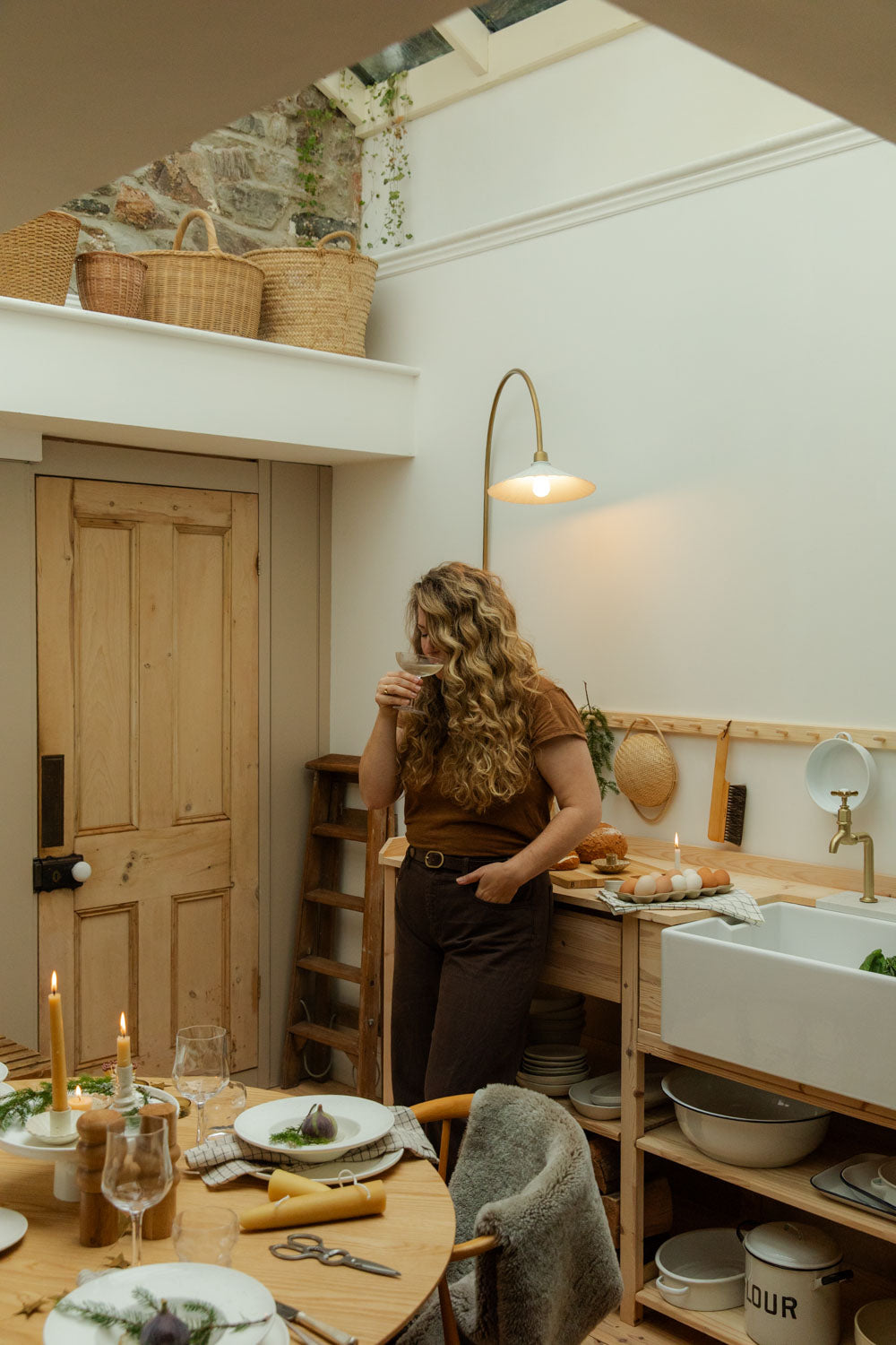 Woman standing in a kitchen with wooden cabinets, sipping a glass of white wine.