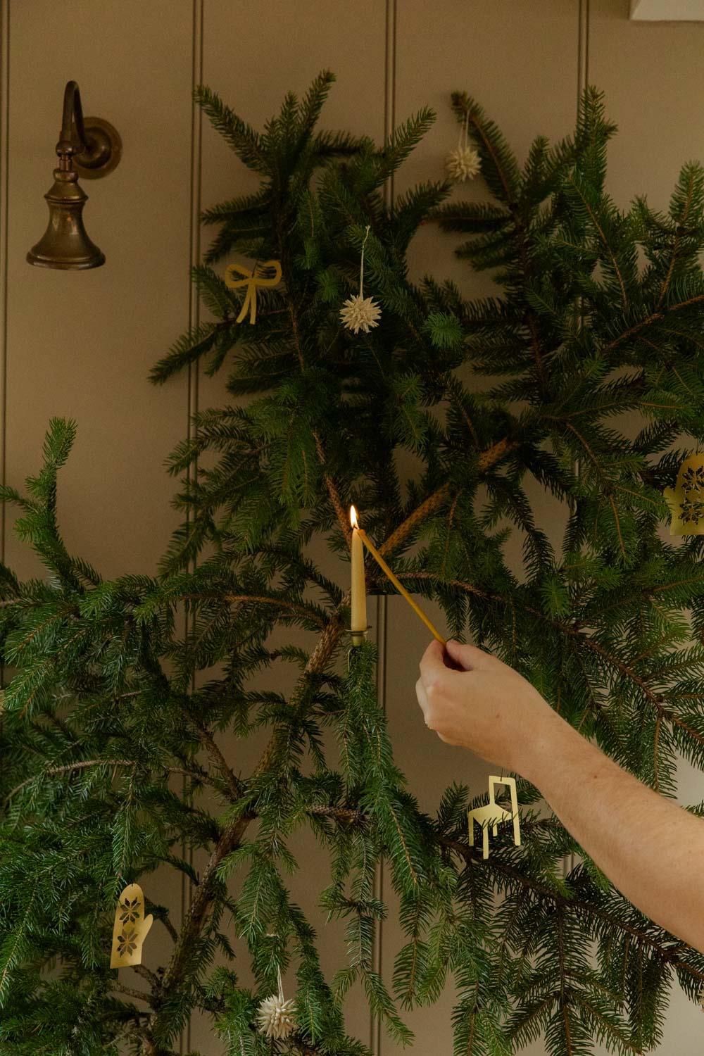 Person lighting a candle on a decorated Christmas tree against a neutral wall.
