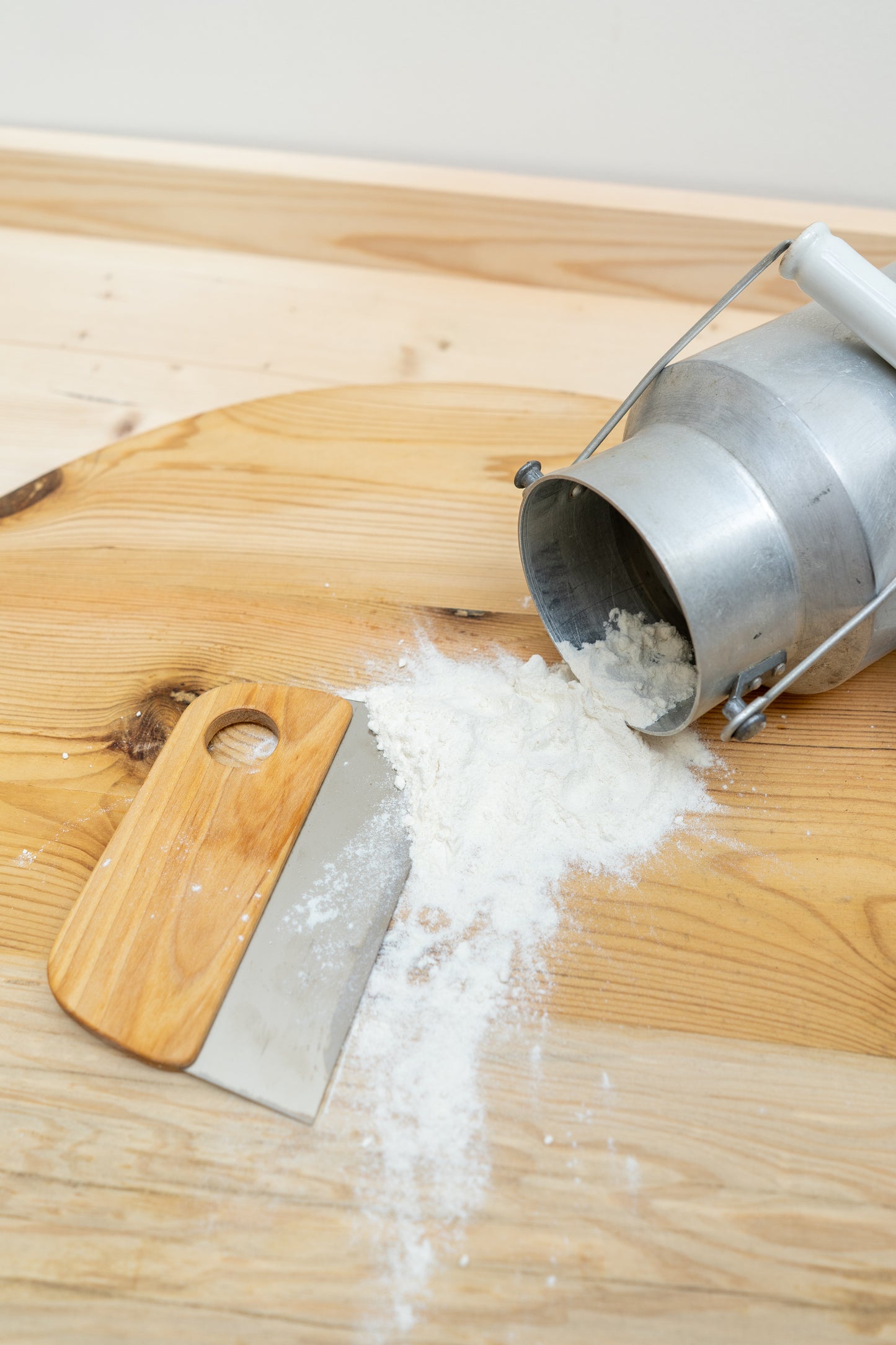 Wooden cutting board with a metal and wood dough scrapper, a pile of flour and metal milk pale on a light background