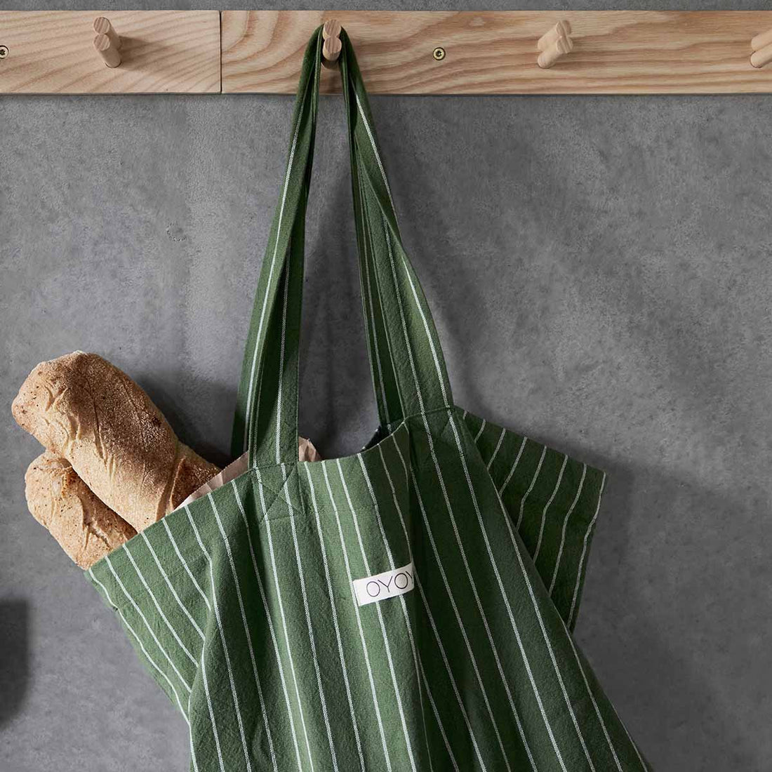 A green striped cotton tote bag hanging on a wooden rail, with a loaf of bread visible in the foreground.