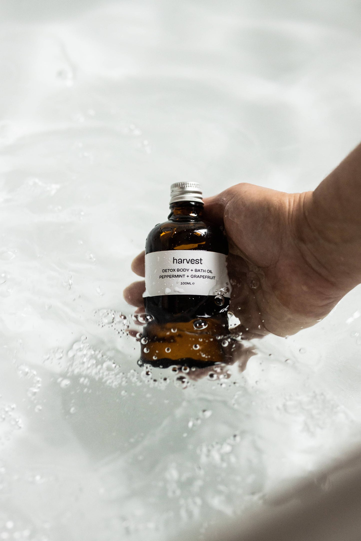 Hand holding Harvest Detox Bath Oil bottle over running bath water
