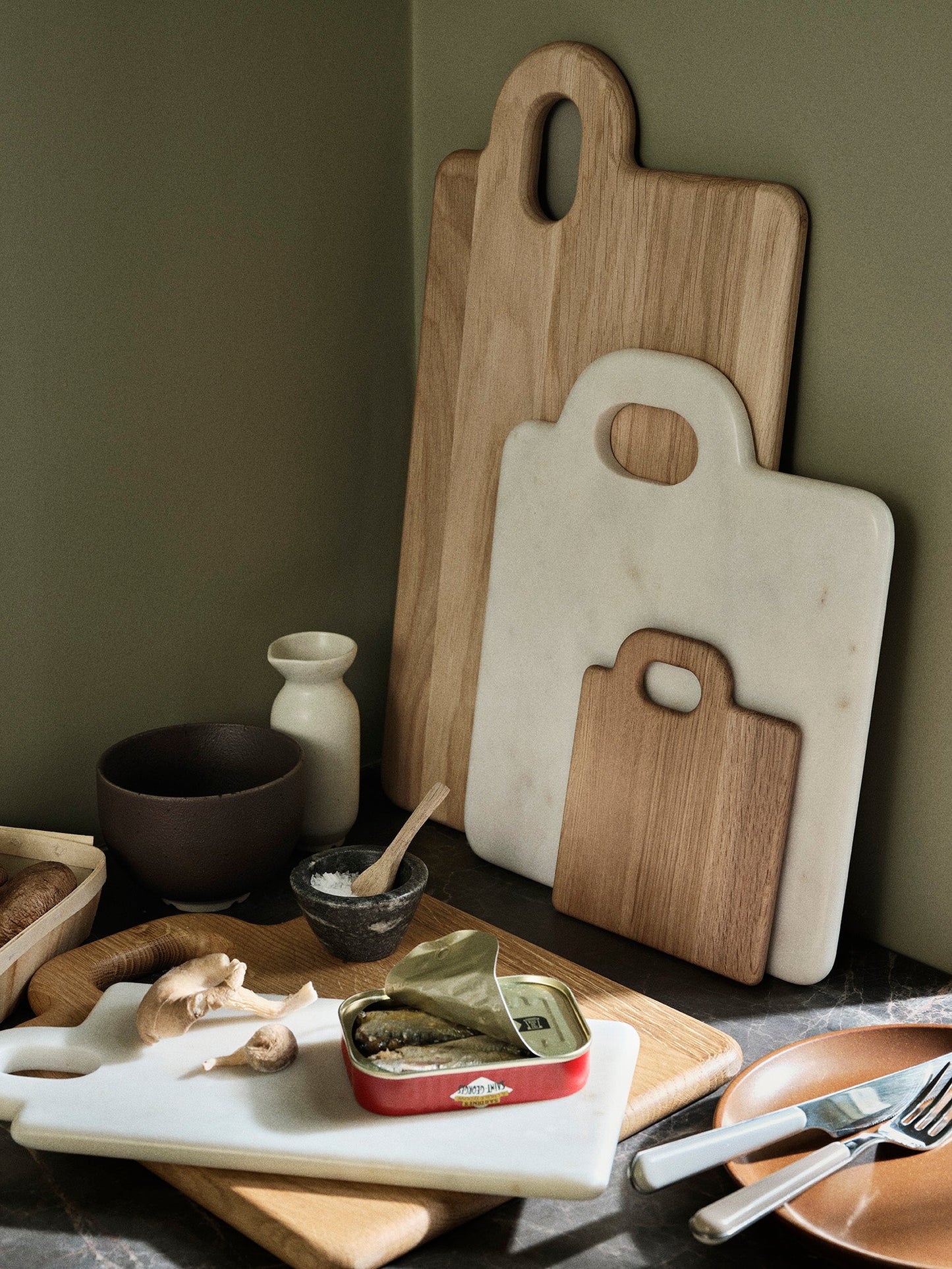chopping boards leaning against an olive green wall, with aesthetically pleasing kitchen mess around.
