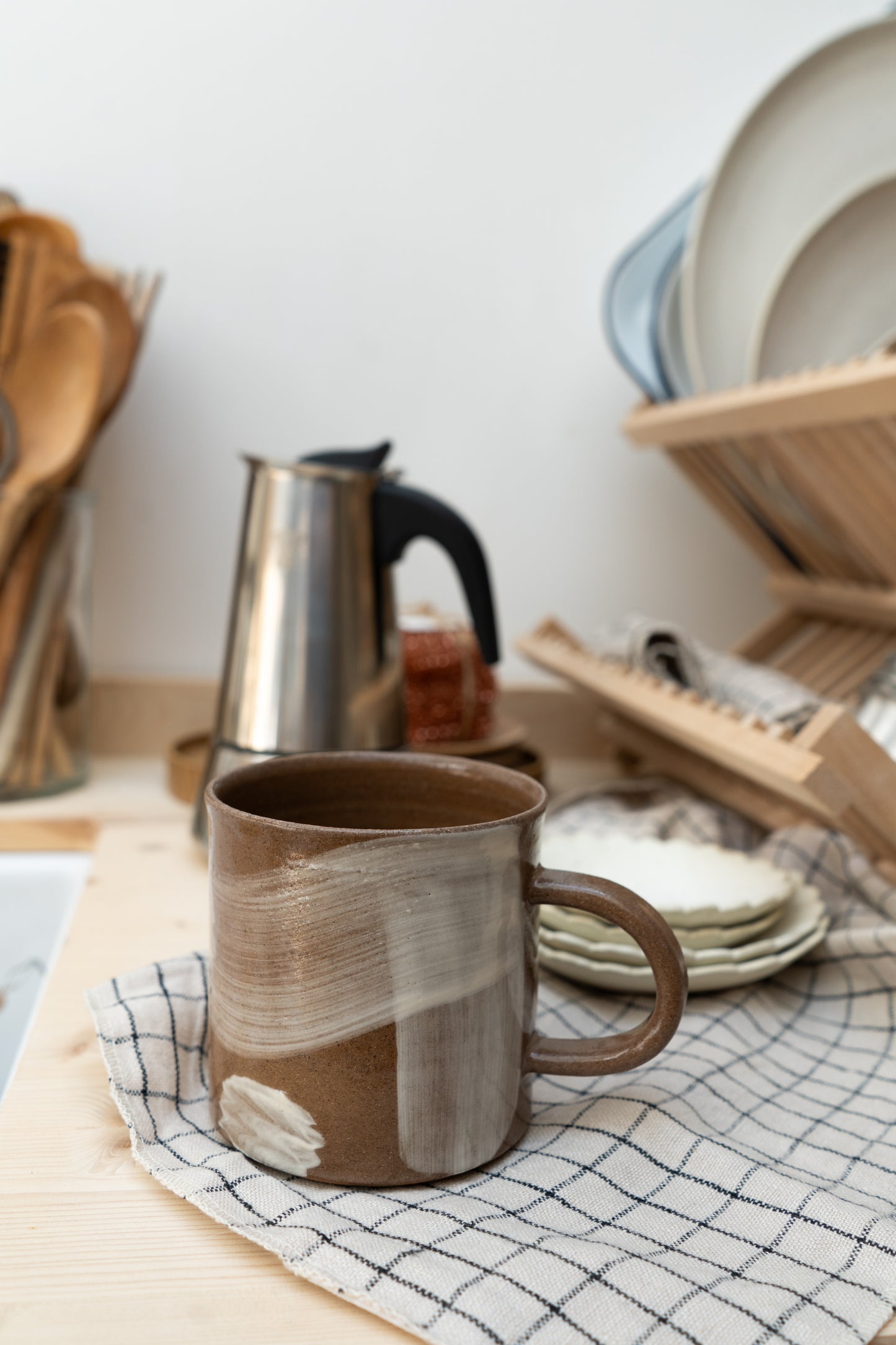 Brown ceramic mug on a checkered towel with a dish rack and kitchen utensils in the background.