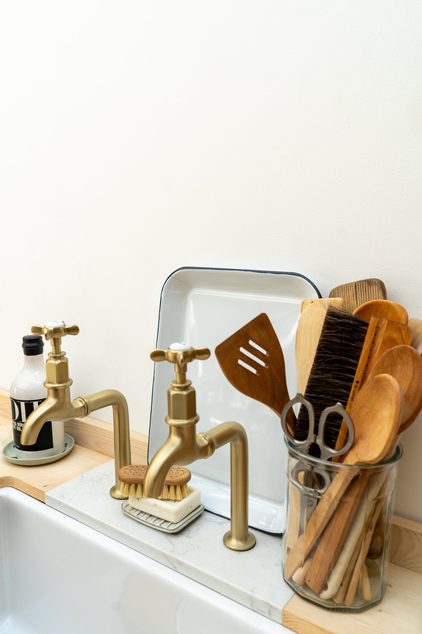 Kitchen sink area with gold faucet, wooden utensils, and cleaning tools on a white countertop.