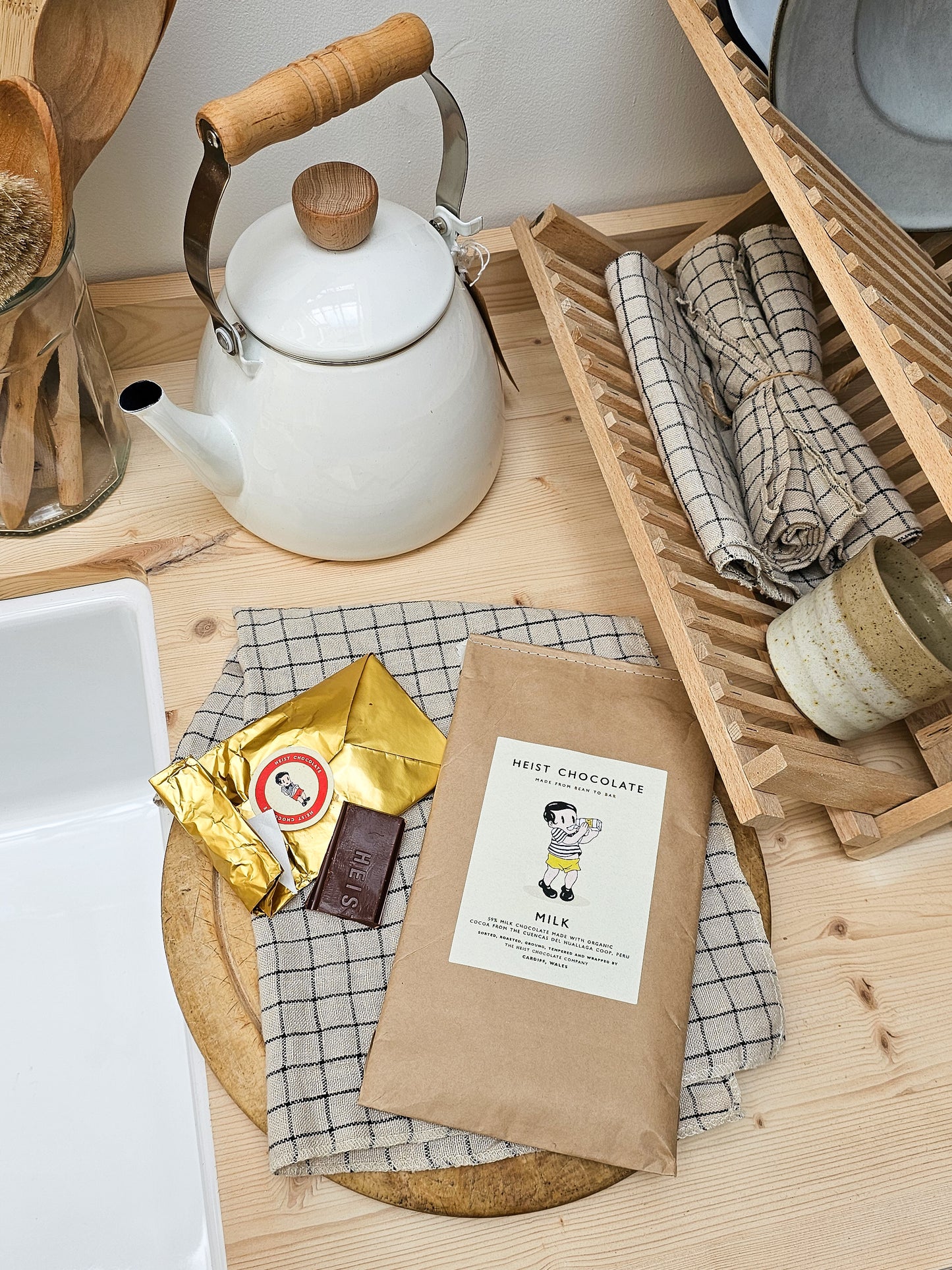 White teapot with wooden handle on a wooden surface with a package of Heist
Chocolate and a bar of chocolate.