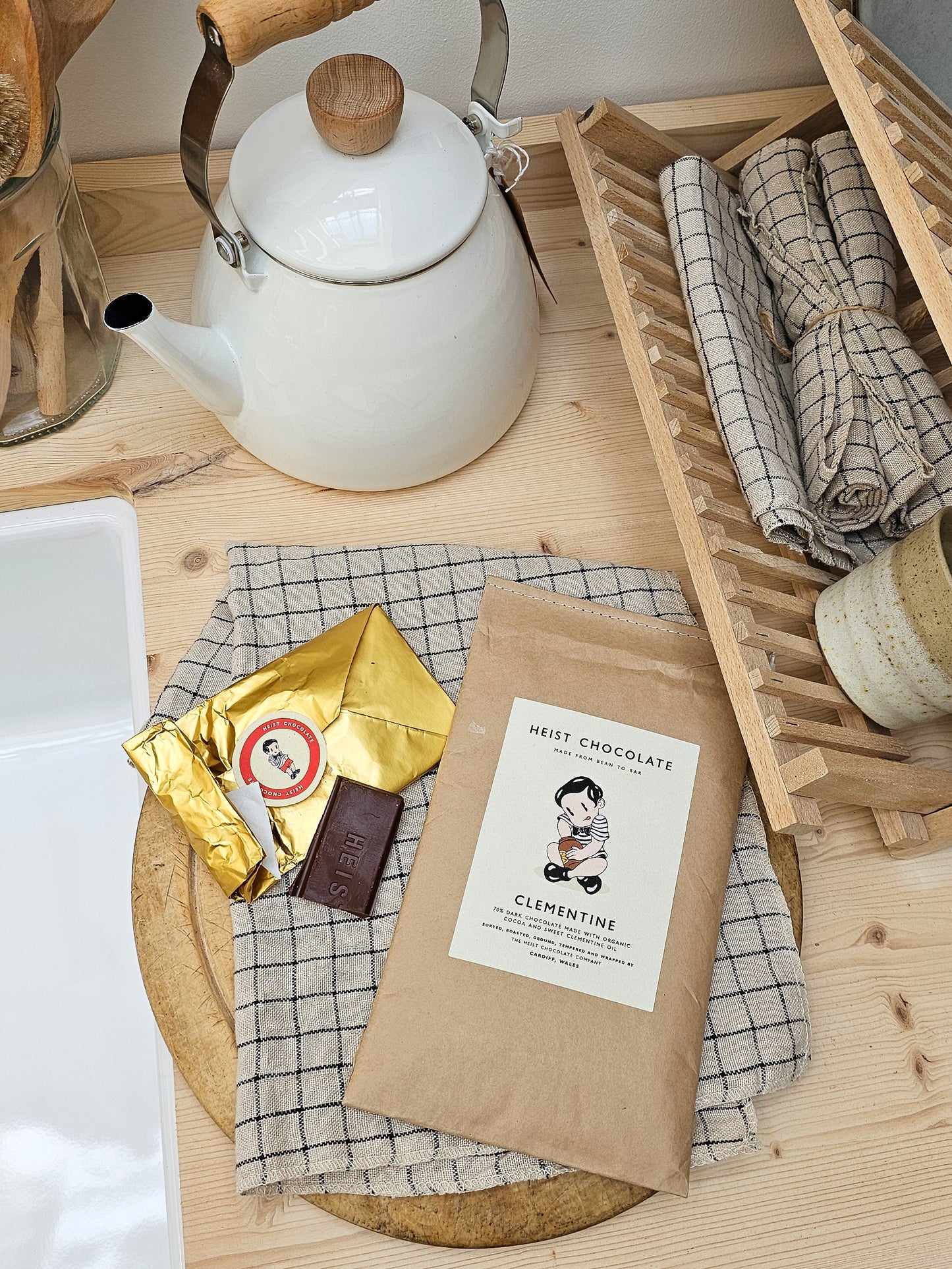 Worktop flatlay view with white teapot, gold chocolate packaging, and chocolate bar on a wooden surface.