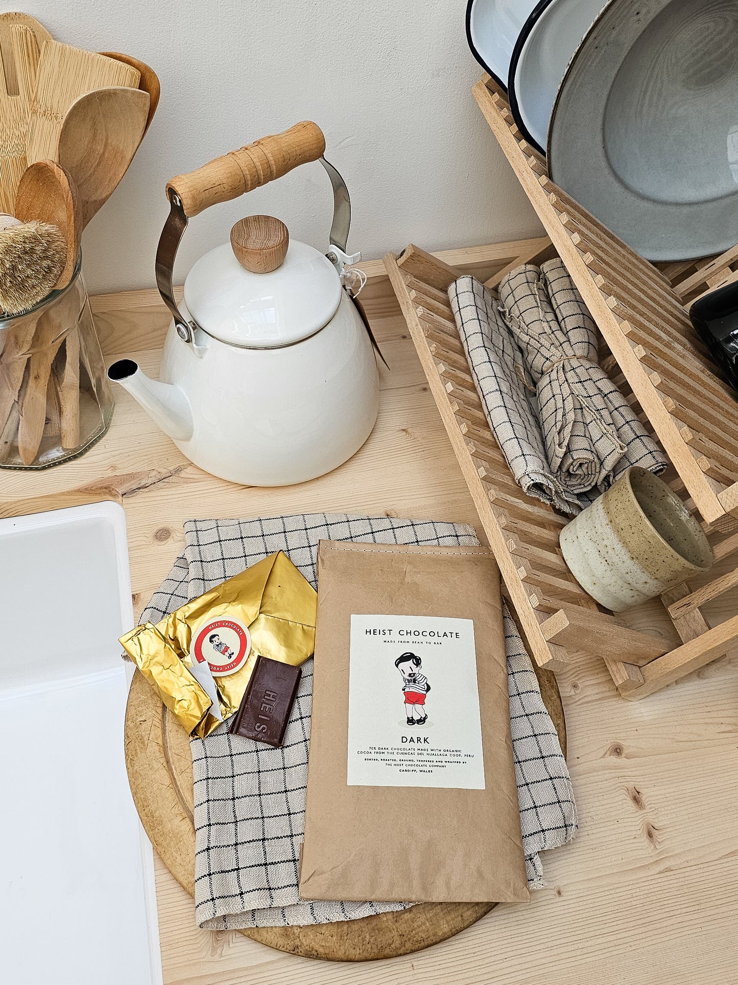 White teapot with wooden handle on a wooden surface with Heist chocolate and kitchen items around