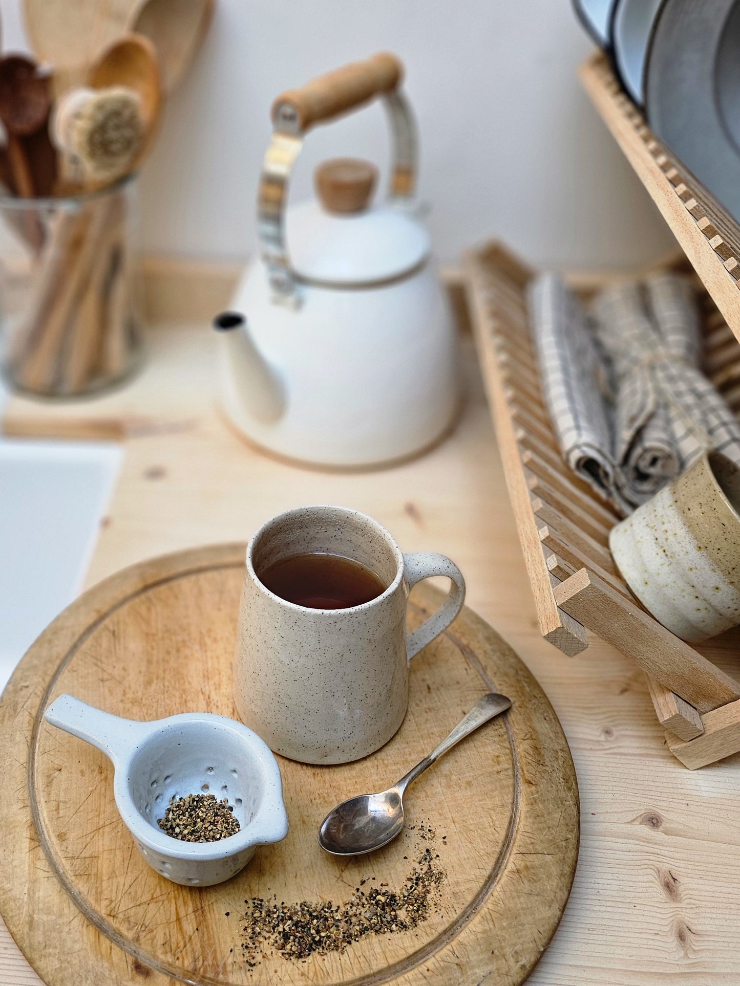 Ceramic mug and tea strainer, on a wooden coaster, surrounded by kitchen items.