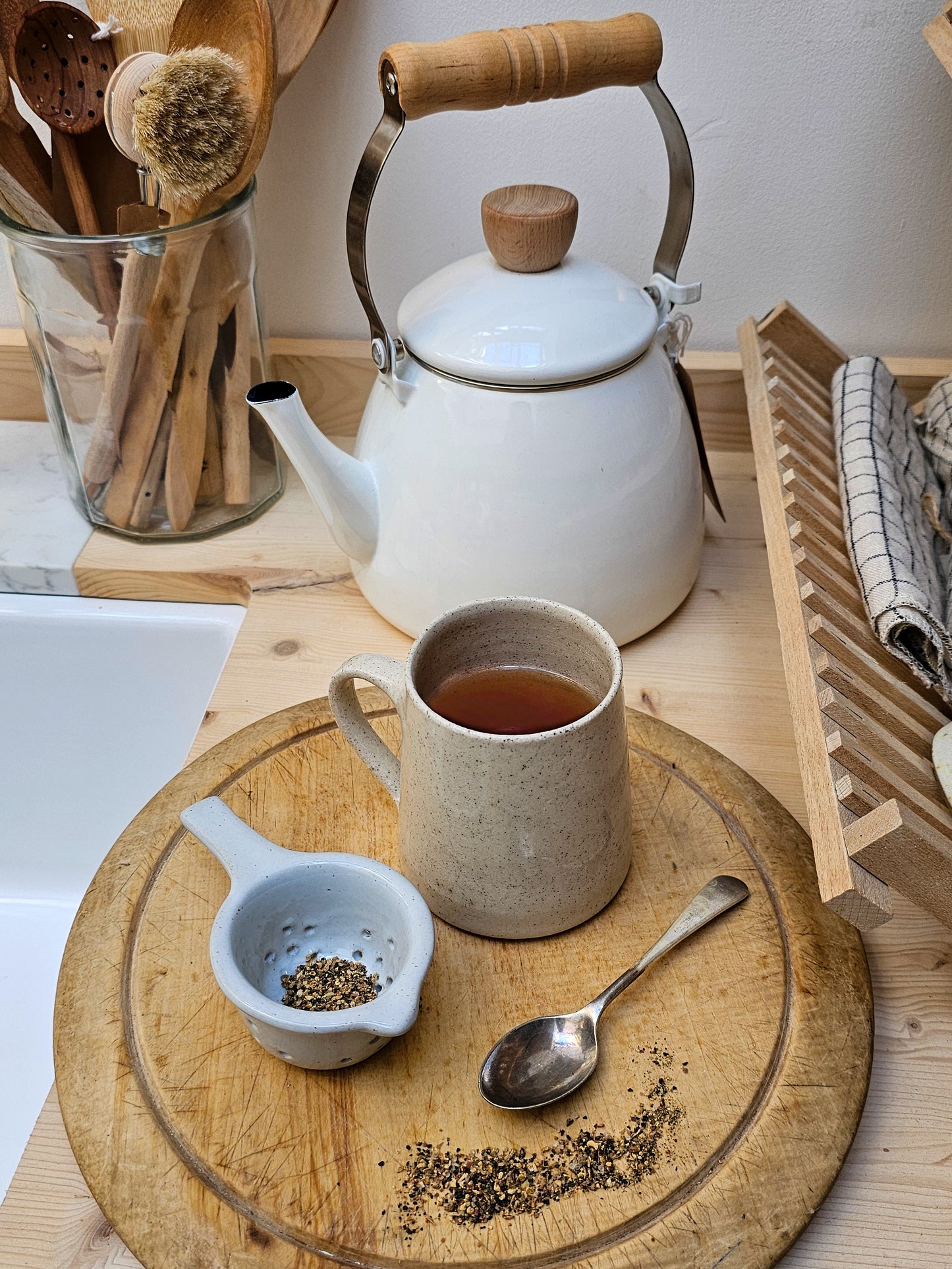 White teapot, mug with tea, wooden spoon, and ceramic tea strainer on a wooden tray.