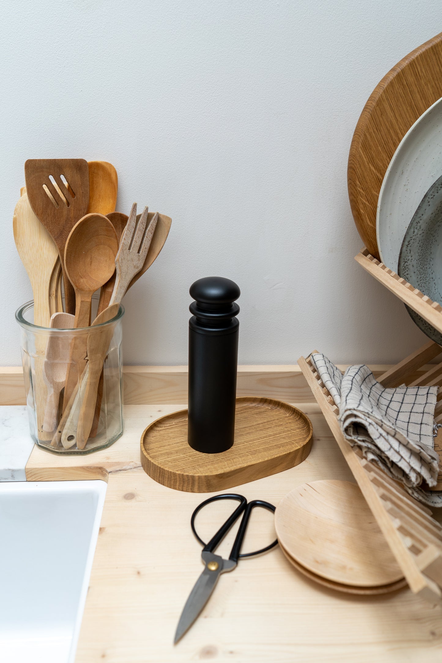 Kitchen utensils, black pepper grinder, and scissors on a wooden countertop with a white wall background.