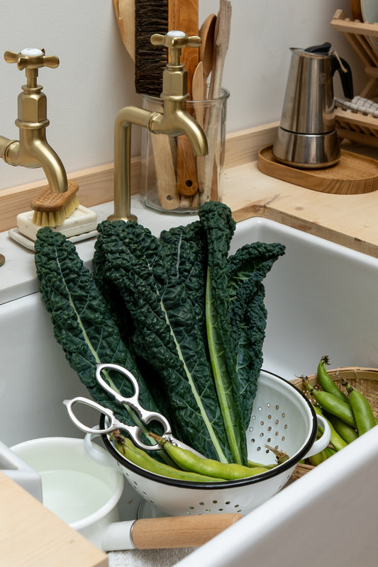 Kale and green beans in a colander in a kitchen sink with utensils and a kettle in the background.
