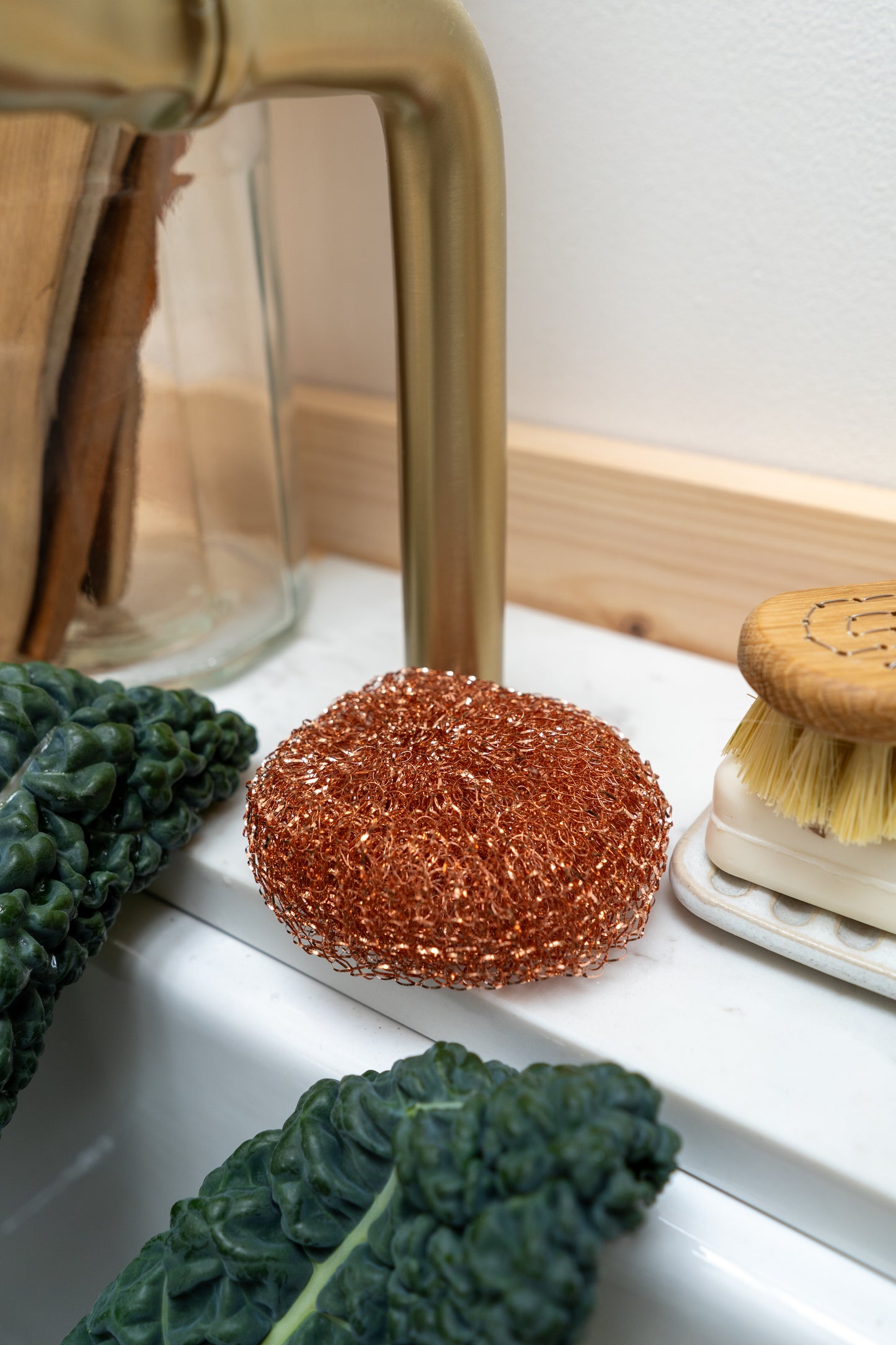 Cleaning products including a copper scrubber, brush, and green leafy vegetable on a white surface.