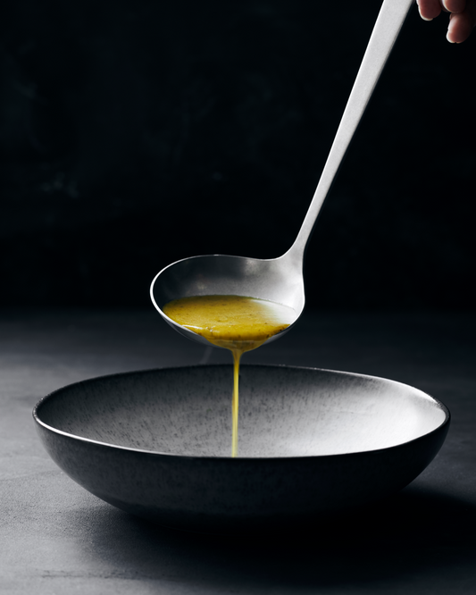 Stainless steel ladle, pouring soup into a back ceramic bowl, against a dark black background.