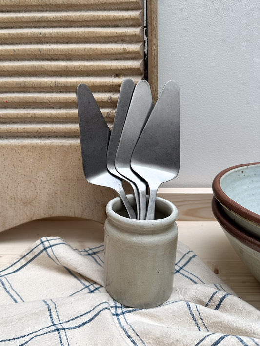 A set of brushed stainless steel cake servers with a spatula-like design, resting in a small ceramic holder, placed on a kitchen towel.