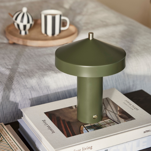 A green LED table lamp with a modern design, placed on a book titled 'Good Living' on a table with a cup and books in the background.