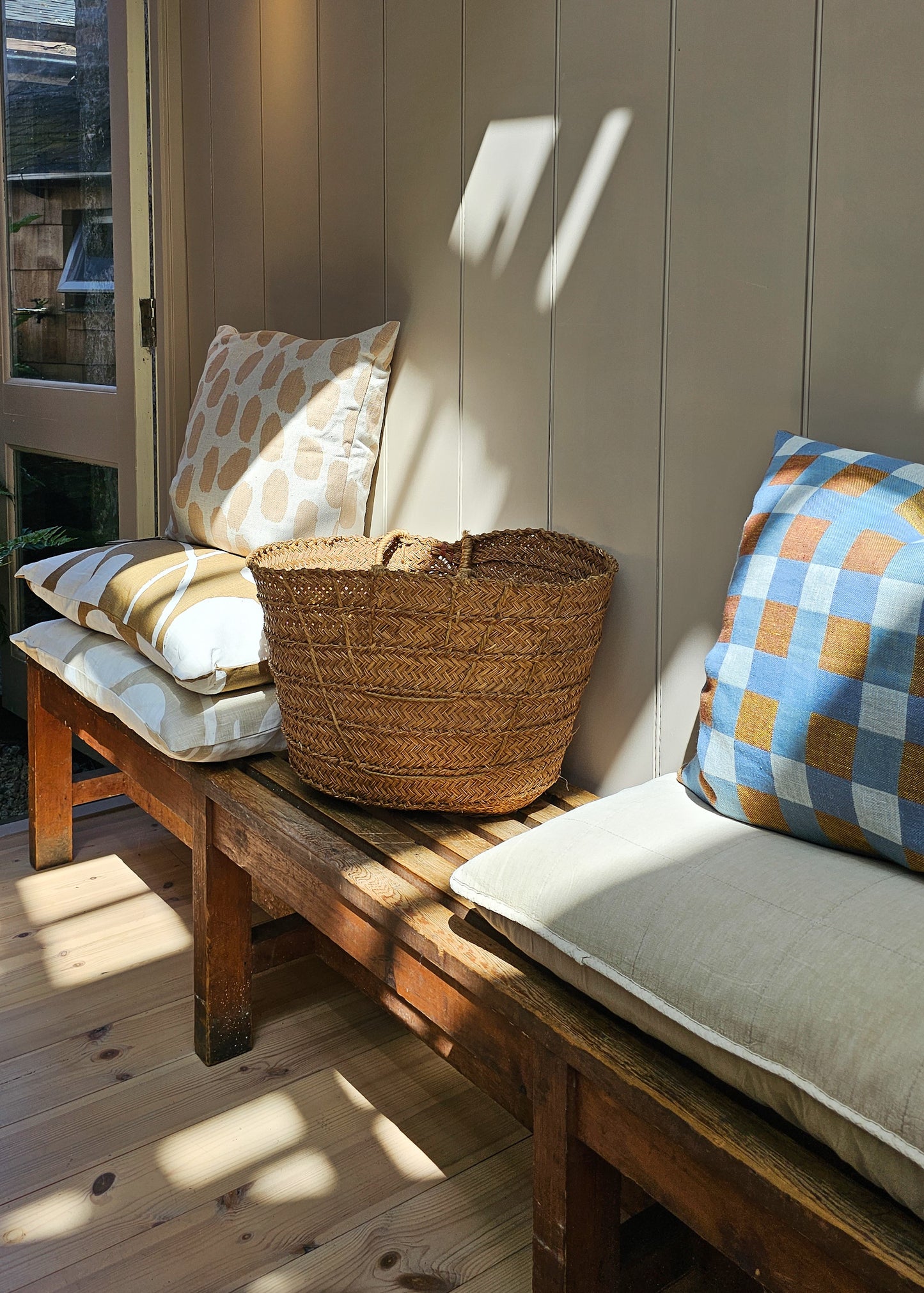 Wooden bench with cushions and a woven basket in a sunlit room.