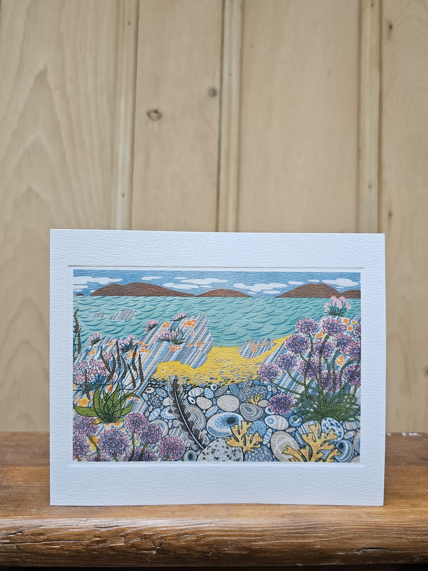 Greeting card reproducing Angie Lewin's print Pebble Shore, showing a richly detailed coastal scene with pebbles, sea thrift, kelp and a feather in the foreground and islands beyond a turquoise sea