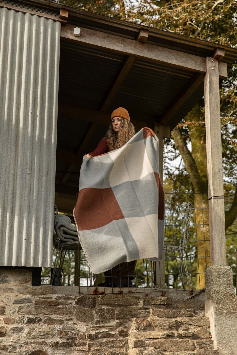 Woman in an orange beanie holding up a Brita Sweden Pop lambswool blanket in an oversized blue, brown and cream block check on a cabin veranda