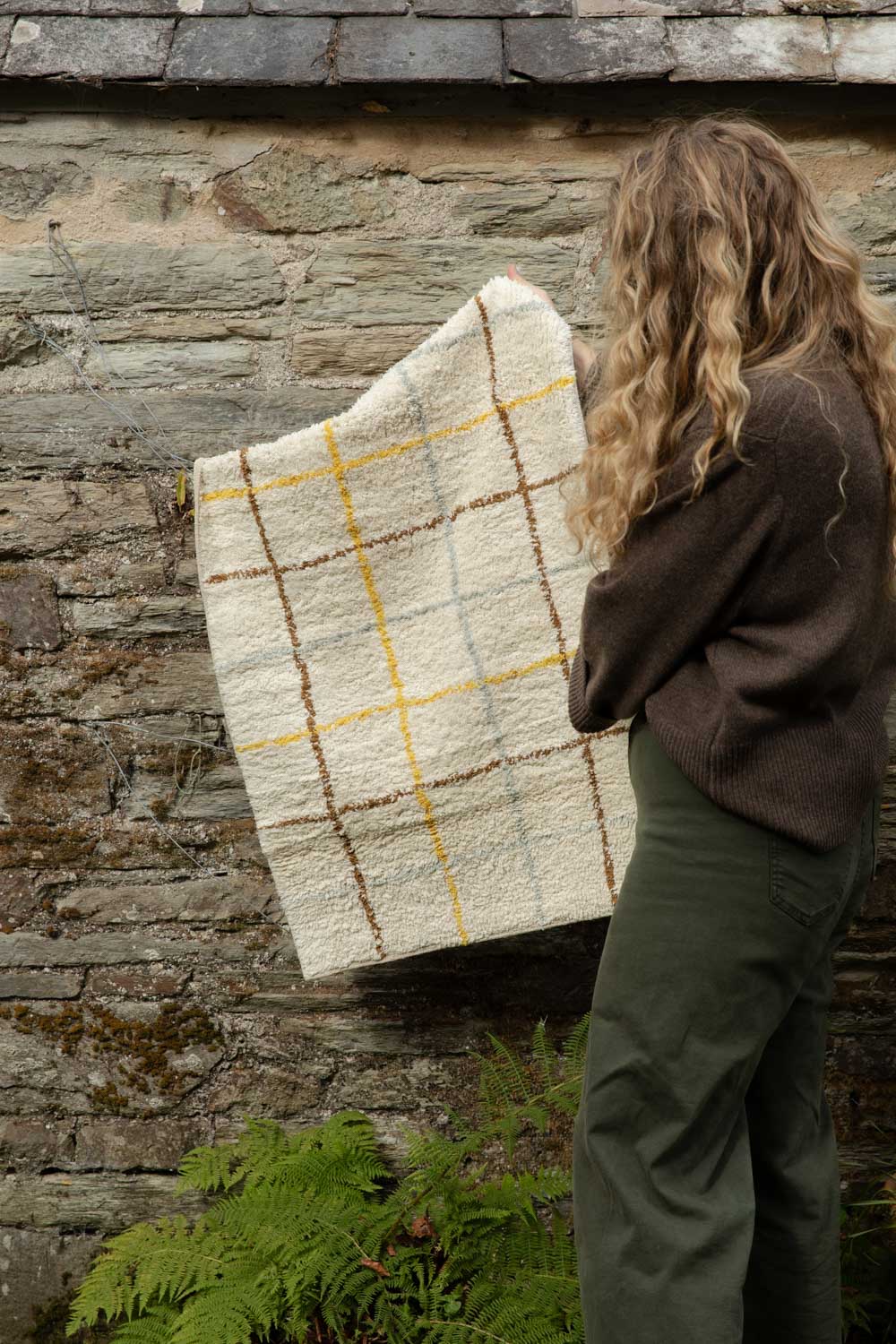 Woman holding up a cream checked mat with yellow and brown lines against a mossy stone wall
