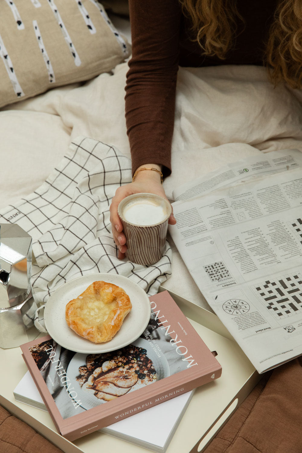 Woman doing a crossword in bed with a Bricka off-white tray holding a Diva stripe mug, pastry, and Bialetti Moka Express.