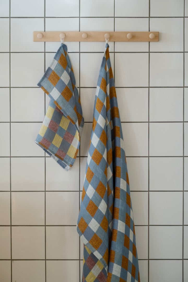A checkered linen tea towel in blue and yellow hanging on a wooden rack against a white tiled wall.