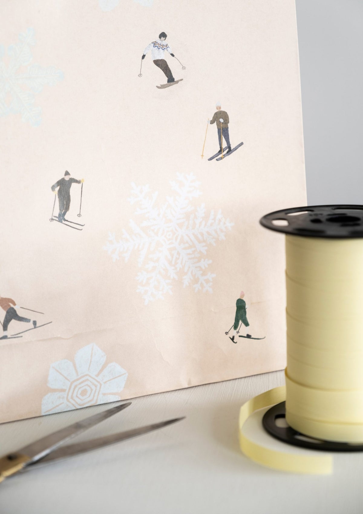 Spool of yellow ribbon in front of gift bag paper featuring skiing illustrations on a white surface.