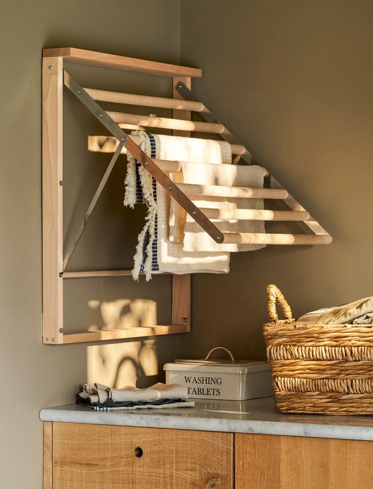 Garden Trading Chalford folding wall dryer in oak with linen tea towels draped over rails, styled above a marble worktop with wicker basket and washing tablets tin