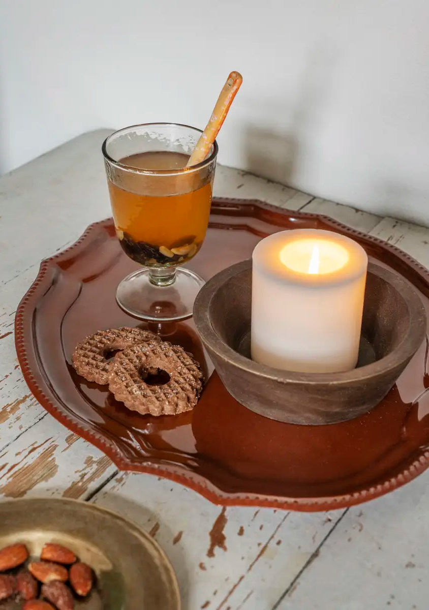 A rustic brown ceramic tray holding a lit white pillar candle in a wooden holder, a glass of amber-colored drink with a cinnamon stick, and two croissants, set on a distressed white wooden table.