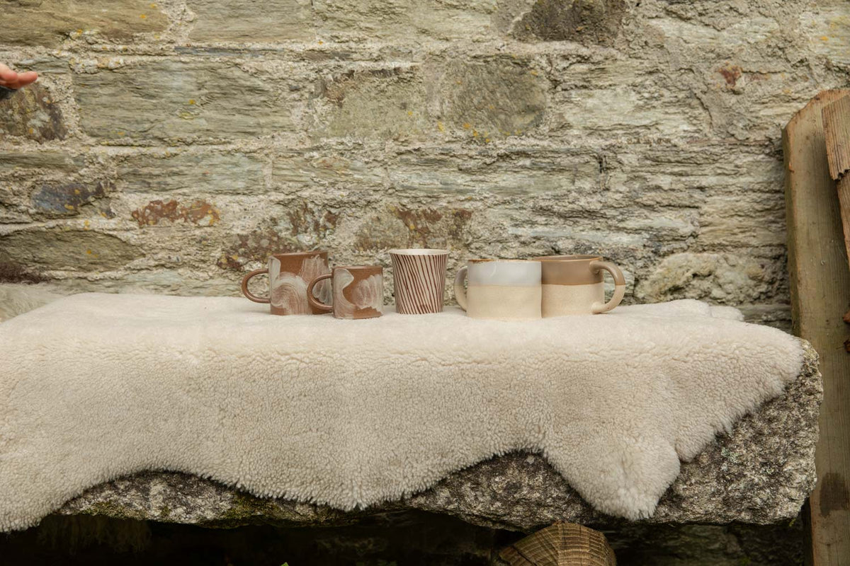 Row of handmade stoneware mugs, brushstroke, stripe and half-glazed, lined up on a cream sheepskin over a granite bench