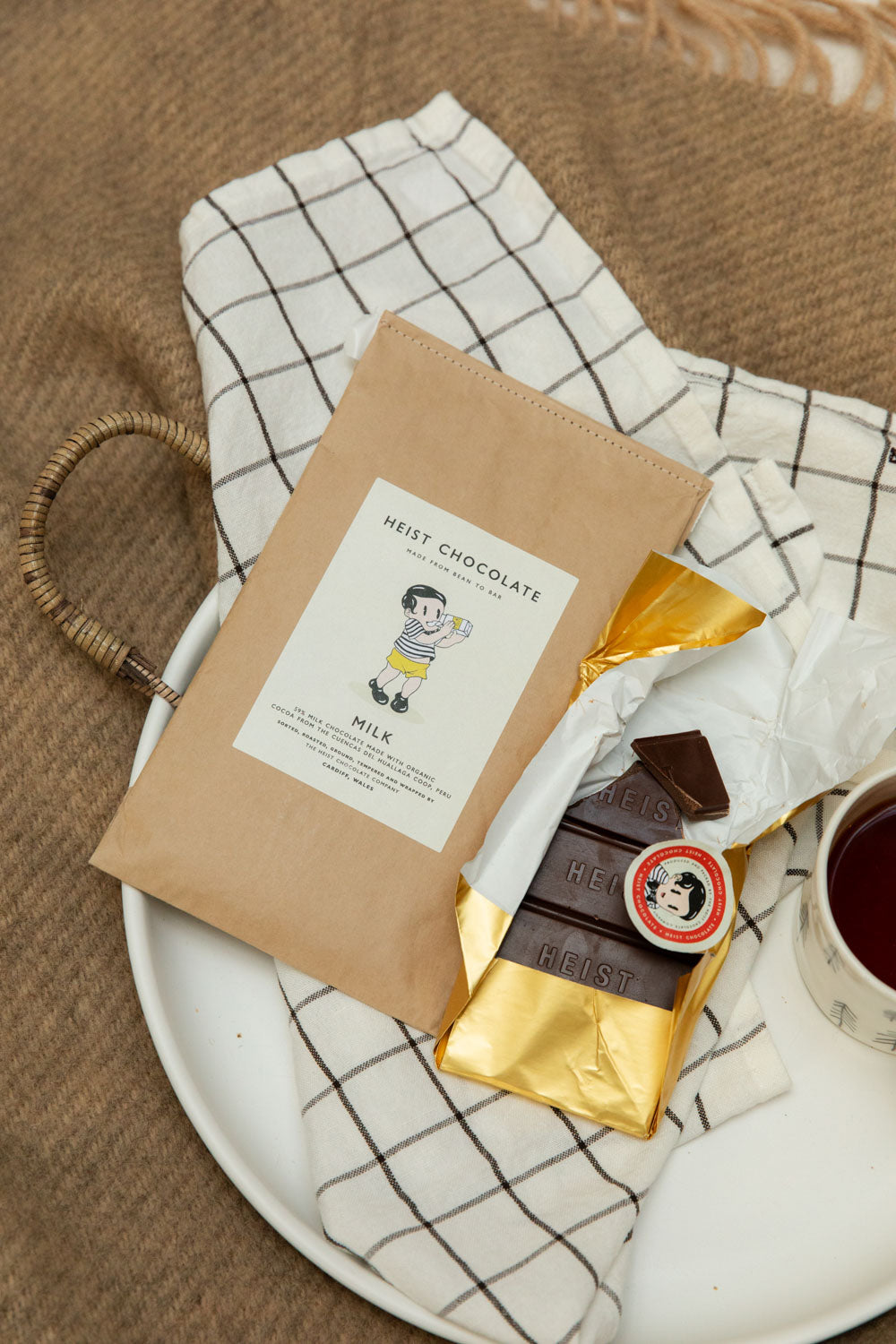 Heist Chocolate Milk bar partially unwrapped in gold foil on a white ceramic tray with a cup of tea and Heist sticker seal.