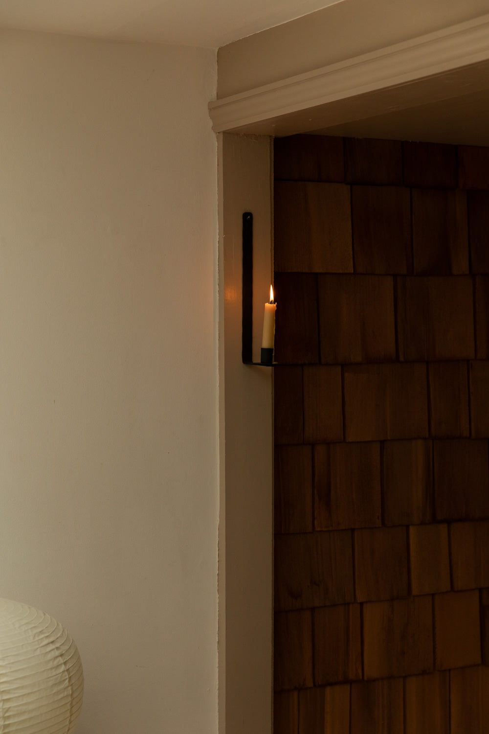 Ib Laursen black iron wall candle sconce with lit taper candle mounted beside a timber clad doorway with a paper lantern lamp visible in the foreground.