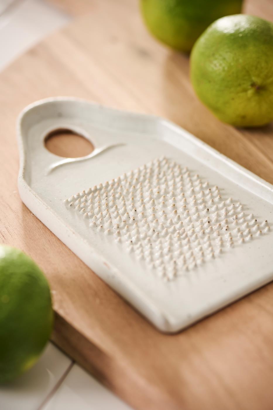 White grater on a wooden surface with green fruits in the background