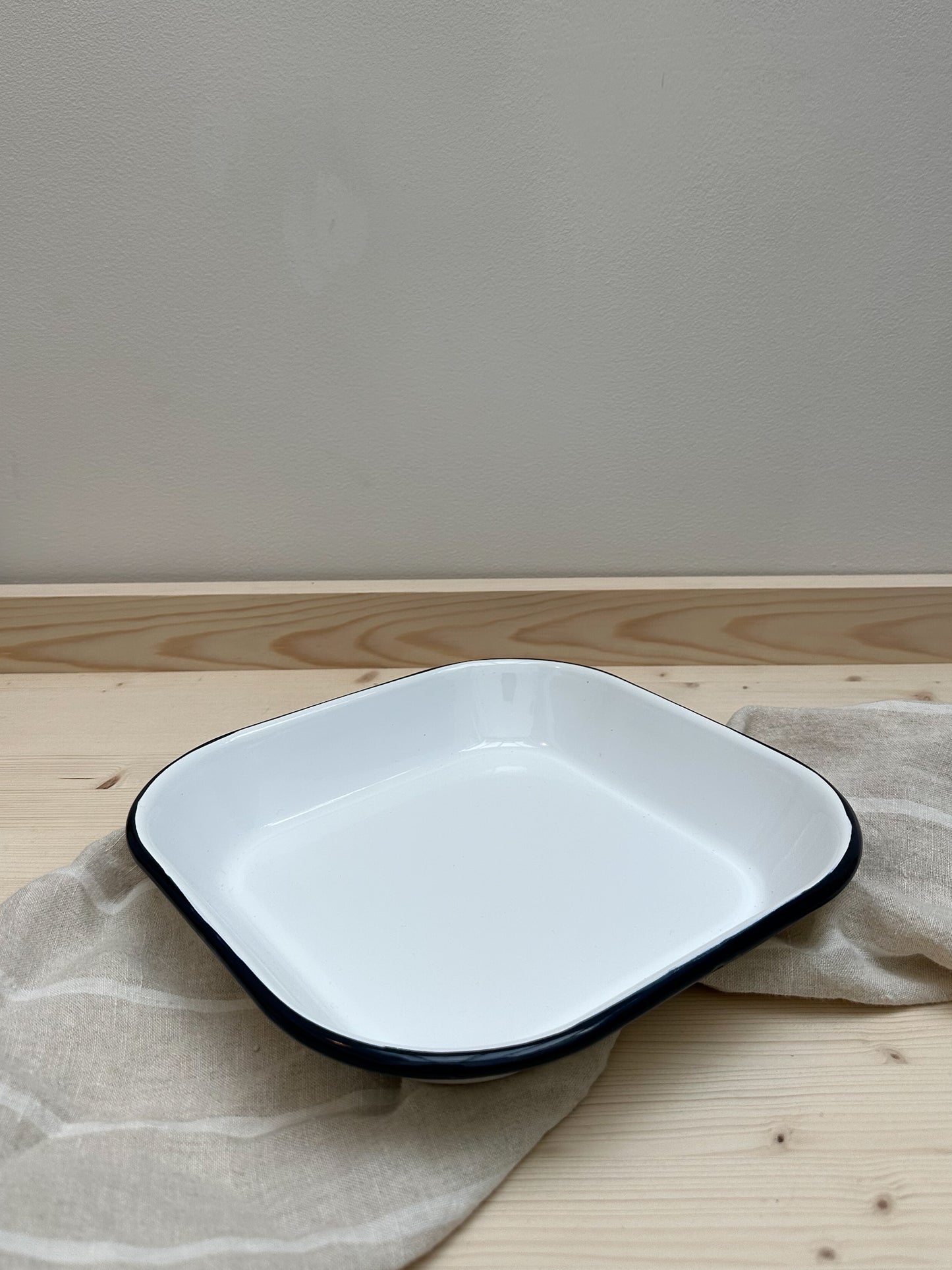 A square enamel baking tray with a white base color and black rim, placed on a wooden surface with a neutral-colored linen cloth.