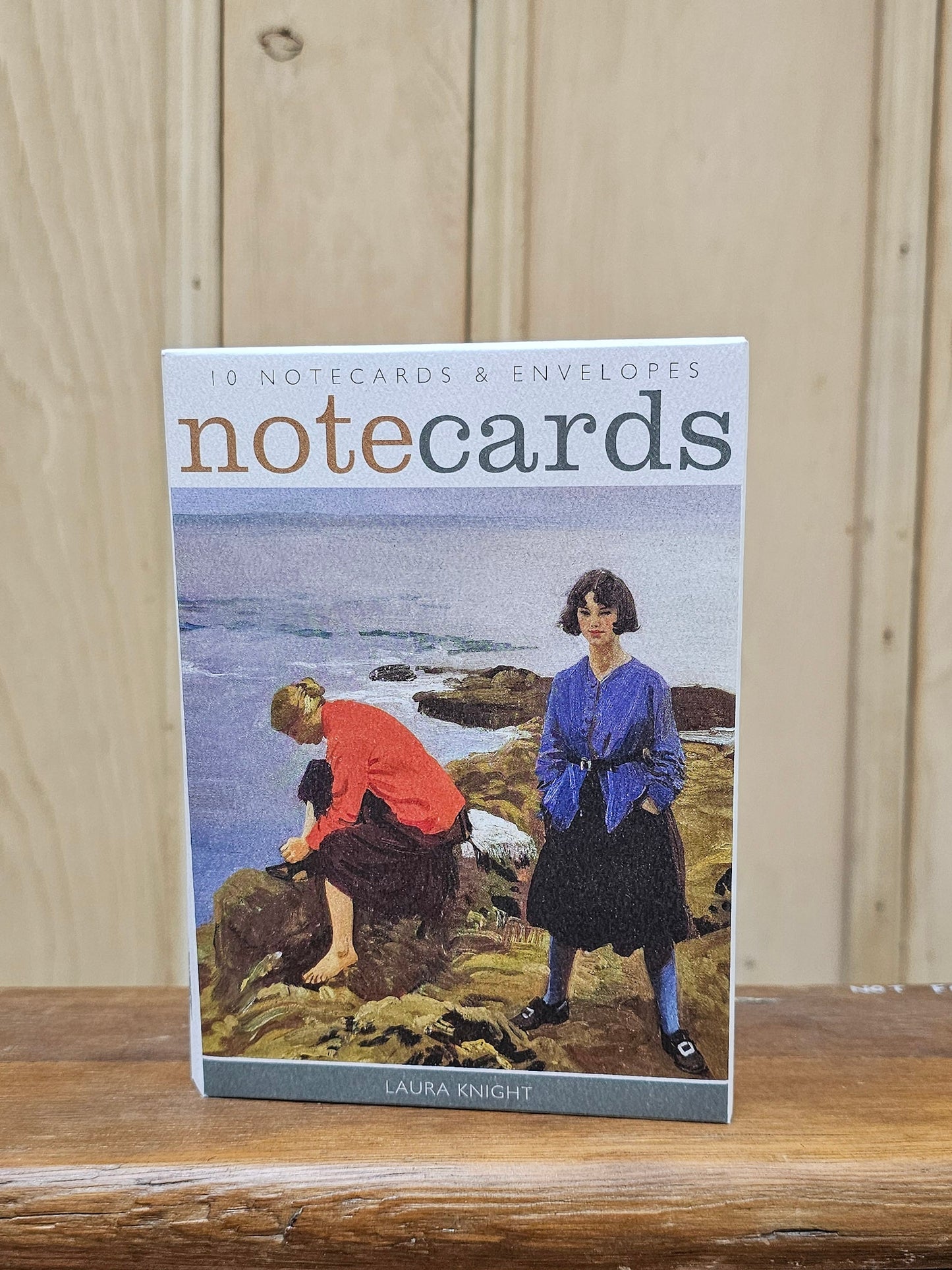 A boxed set of notecards and envelopes titled "notecards" by Laura Knight, standing upright on a wooden surface against a light wood paneled background. The front cover shows an illustration of two women by the ocean.