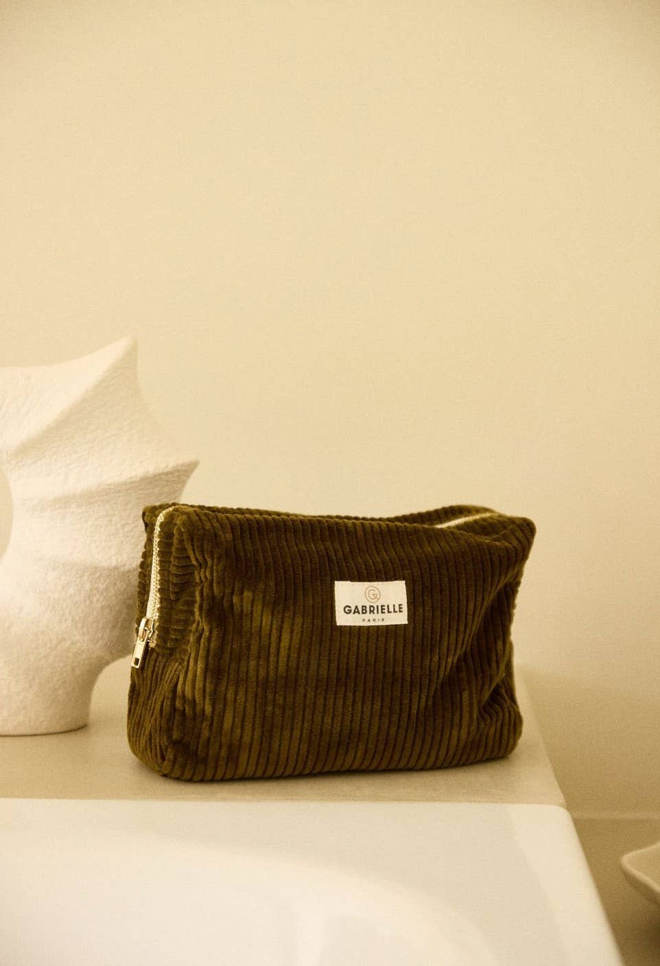 Olive corduroy pouch with a Gabrielle Paris label on a white surface against a beige wall.