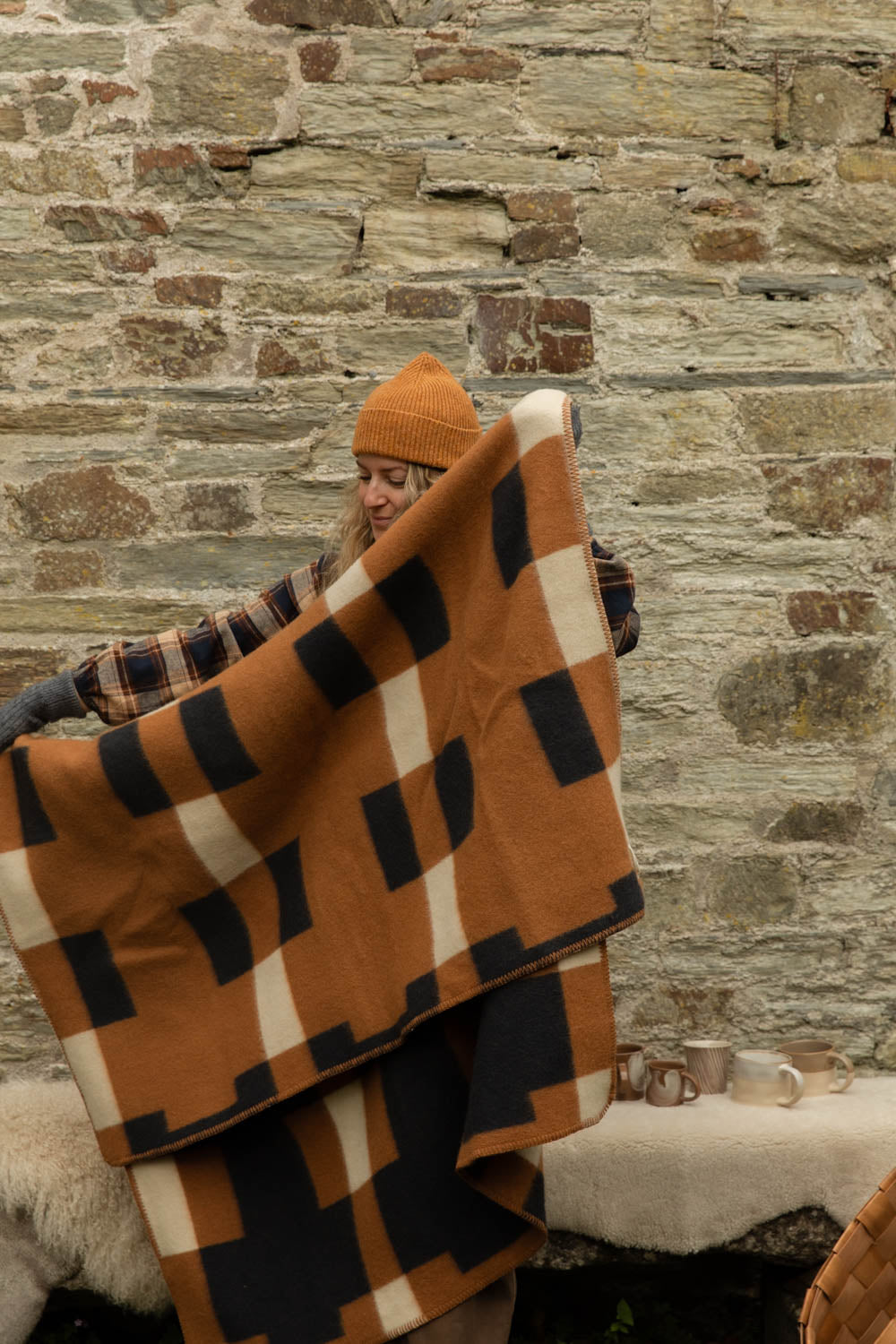 Woman in a mustard ribbed beanie holding open a lambswool blanket in a bold orange, black and cream block check against a stone wall
