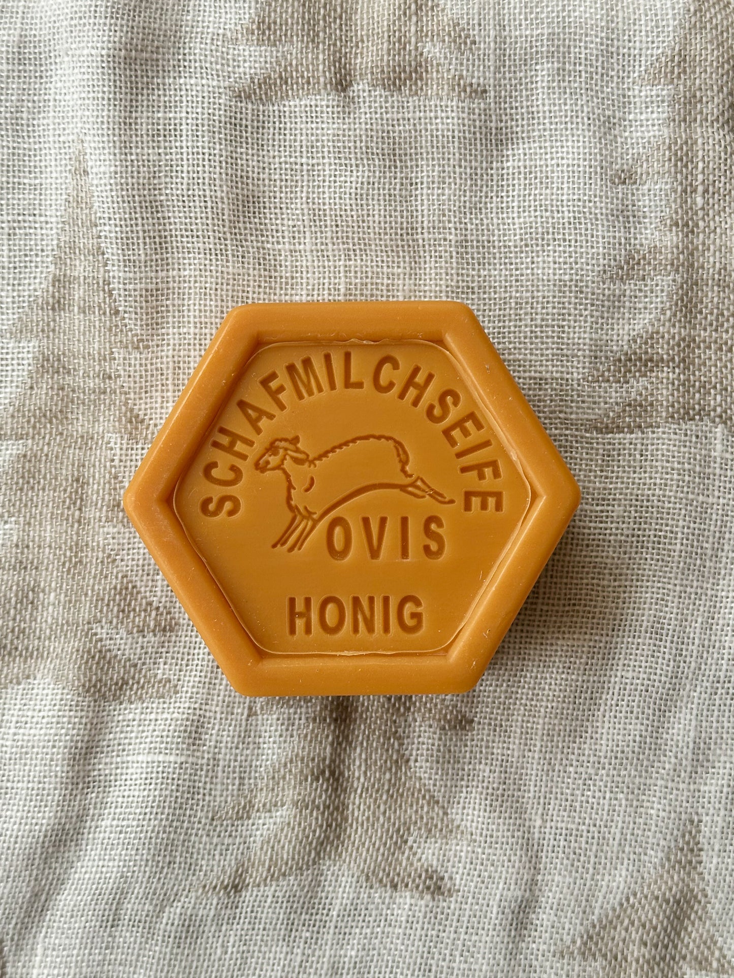 A hexagonal bar of soap with a solid yellow color and text on it, placed on a textured fabric surface.