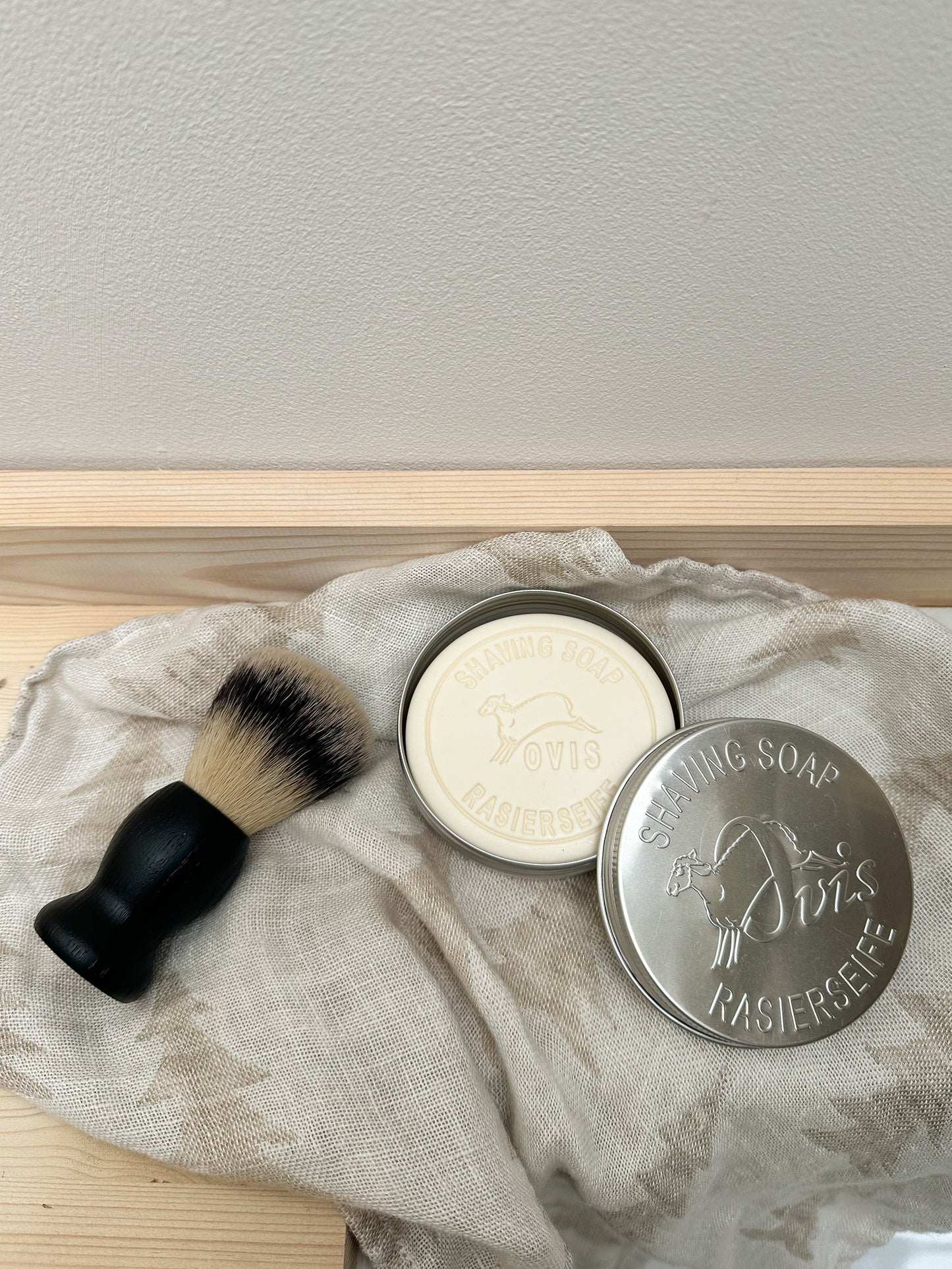 Shaving brush and shaving soap bar in an open silver tin, on a textured cloth with a neutral background