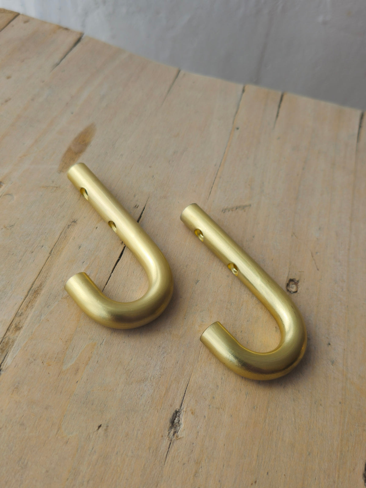 Two brass metal J-hooks with pre-drilled holes, resting on a light-colored wooden surface.
