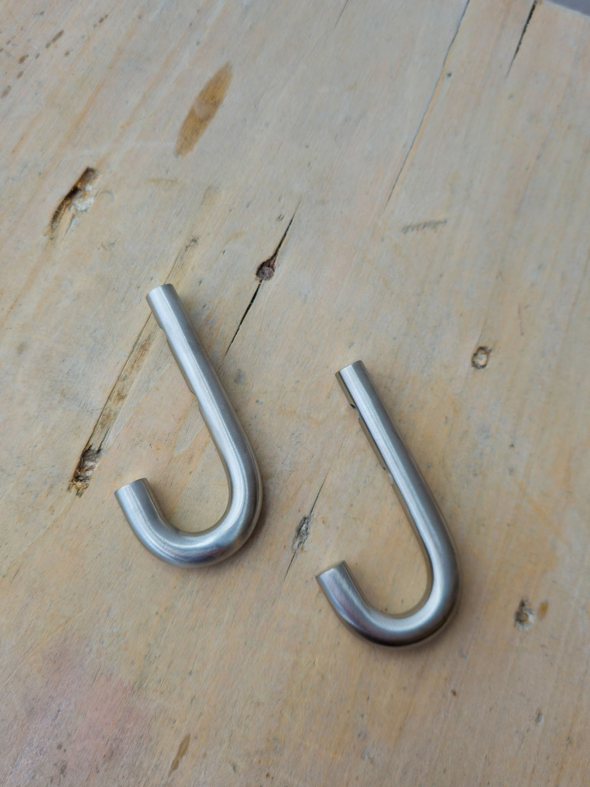 Two silver metal J-hooks lying on a light-colored wooden surface.