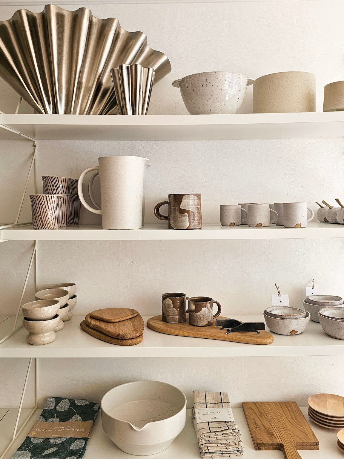 White metal shelving displaying a collection of ceramic tableware, including bowls, mugs, pitchers, and serving dishes in neutral beige, white, and brown tones.