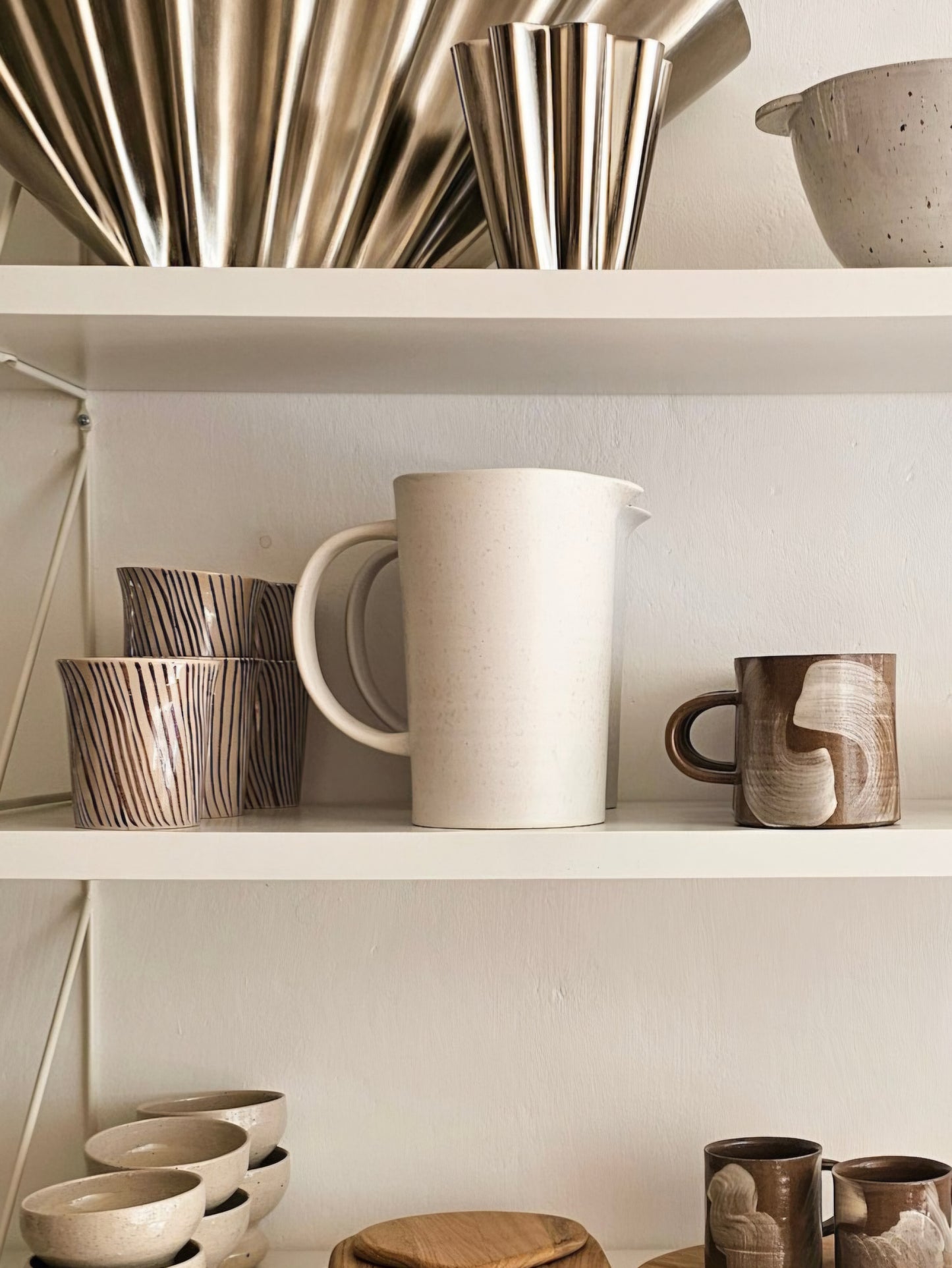 A styled interior shelf display featuring ceramic and metallic homeware, including a large off-white pitcher, patterned cups, bowls, and brushed metal vessels against a warm neutral wall.