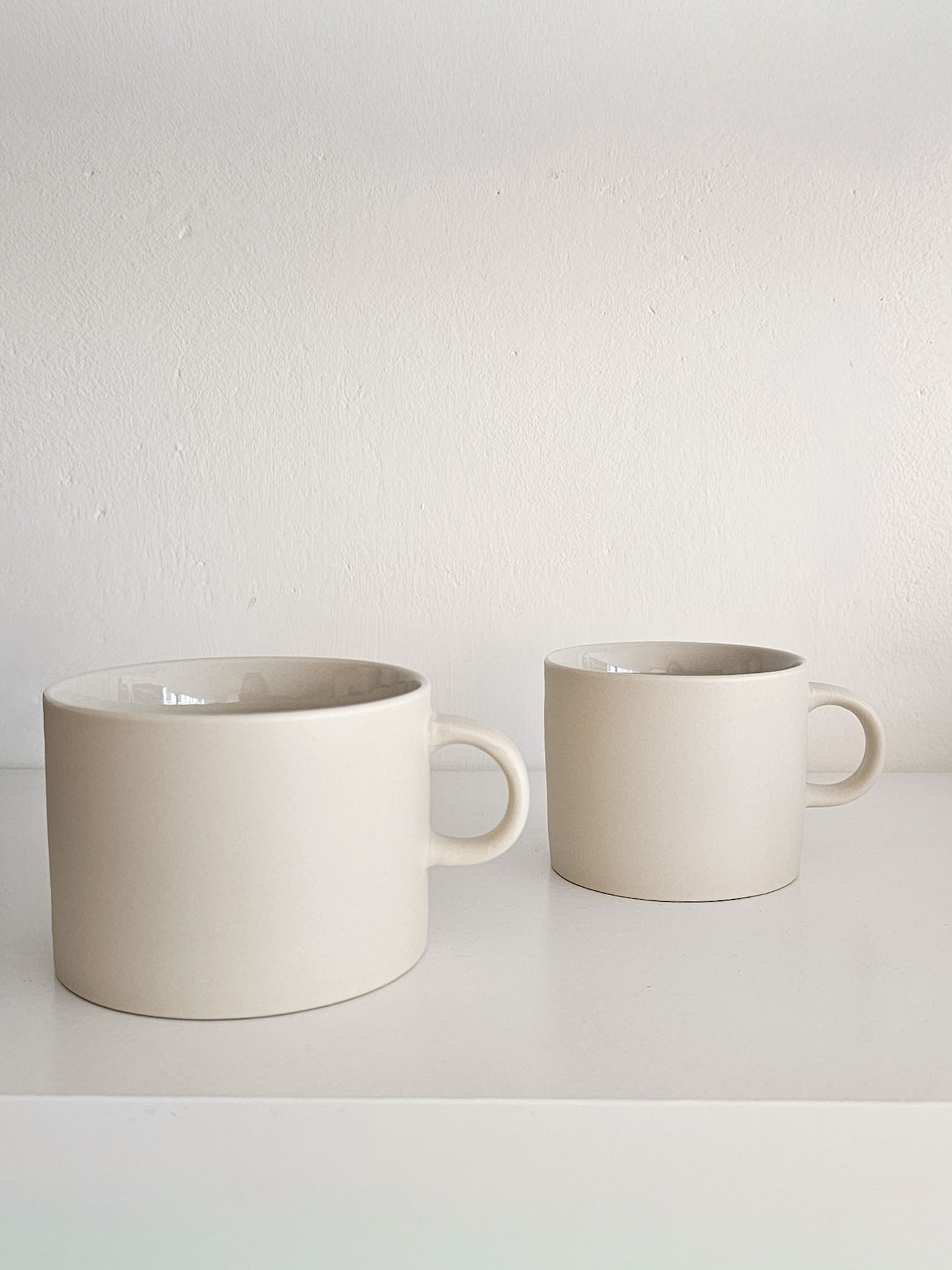 Two matte ceramic mugs in a warm off-white or cream color, displayed on a white shelf against a textured white wall.