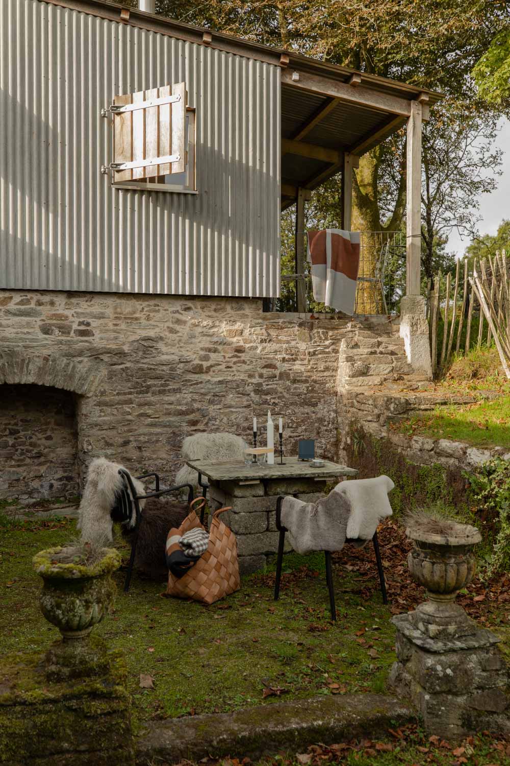 Autumn garden table beside a tin-roofed cabin, with long-haired Tibetan sheepskins draped over black chairs and a woven basket on the grass
