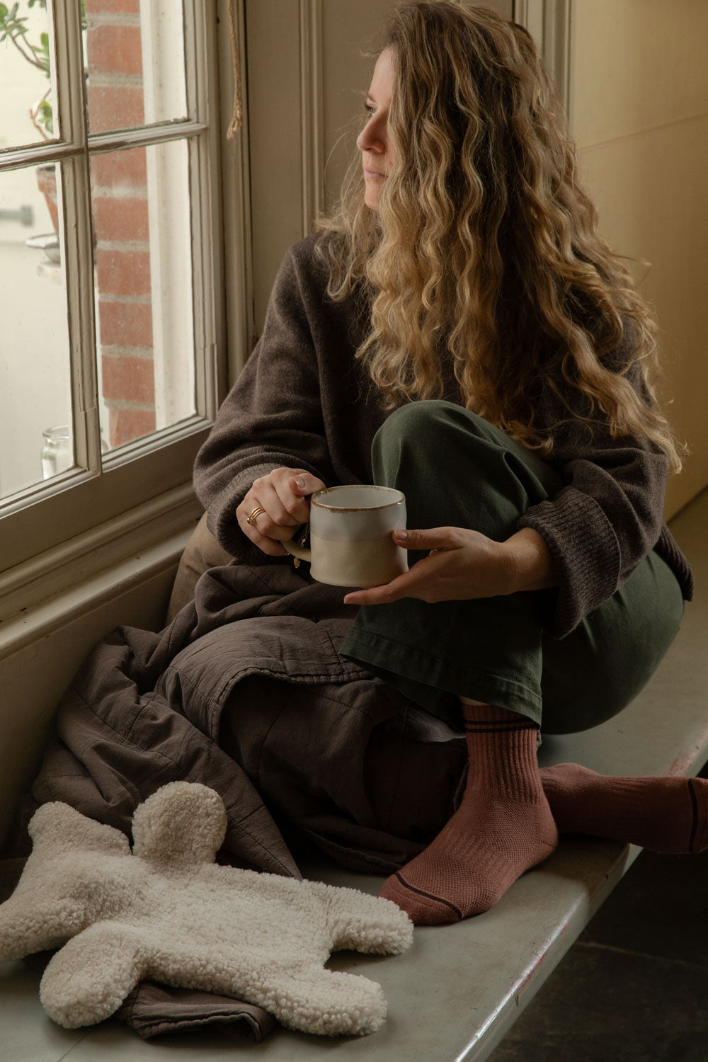 Woman with a mug gazing out of the window, a cream teddy sheepskin hot water bottle on the seat