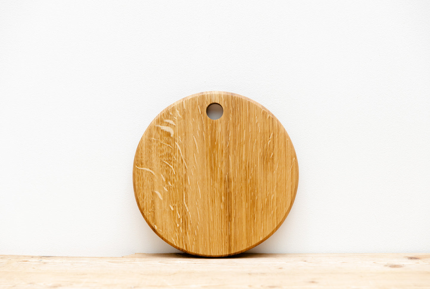A round, English oak wood board with a hole at the top, placed on a wooden surface, leaning against a white wall.