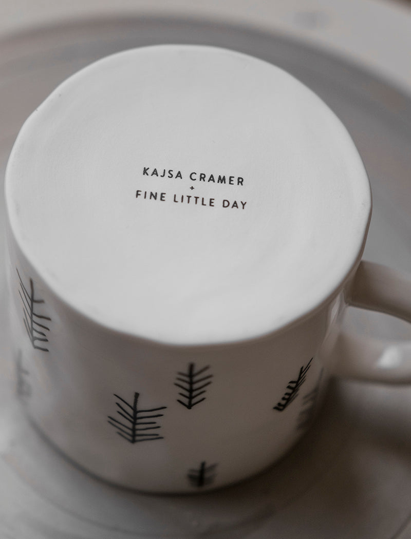 The bottom of a white porcelain cup with a pine tree motif pattern. The writing on the bottom of the cup says Kajsa Cramer + Fine Little Day.