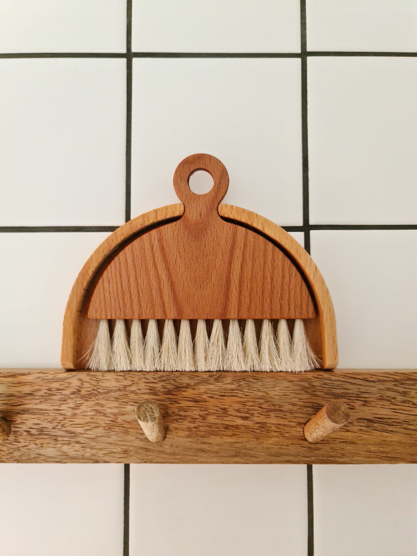 A wooden table brush with a matching dustpan, placed on top of a peg rail against a tiled wool.